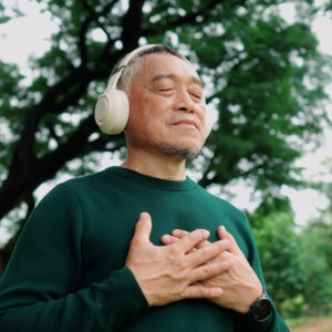 Elderly Asian man meditating under a tree with wireless headphones, calm and refreshed.